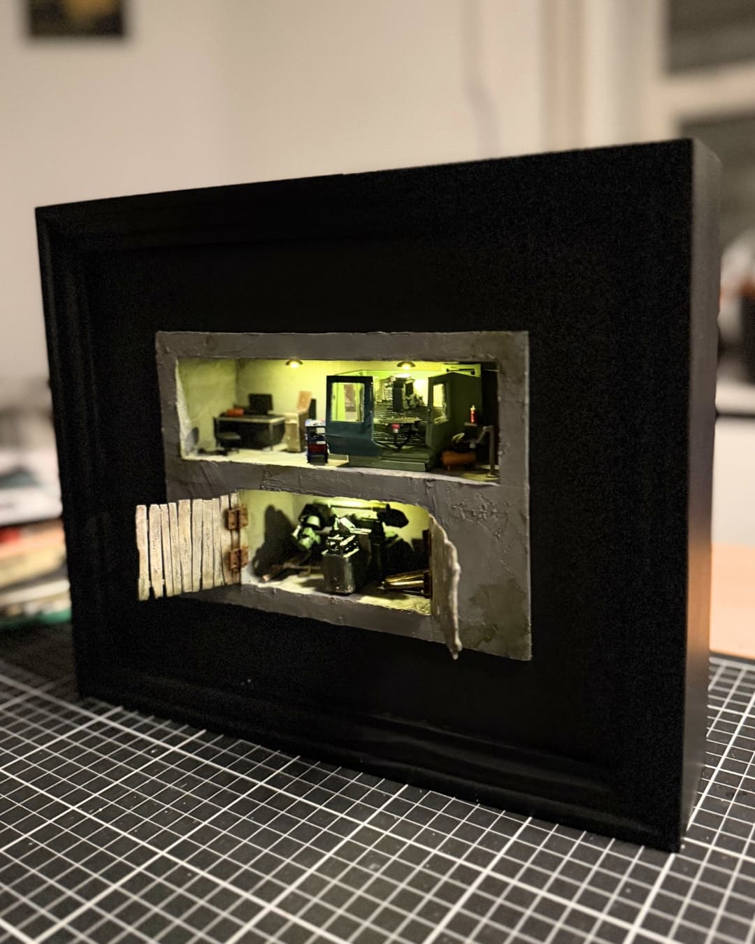 Black framed diorama with a lit two-level workshop behind a broken wall