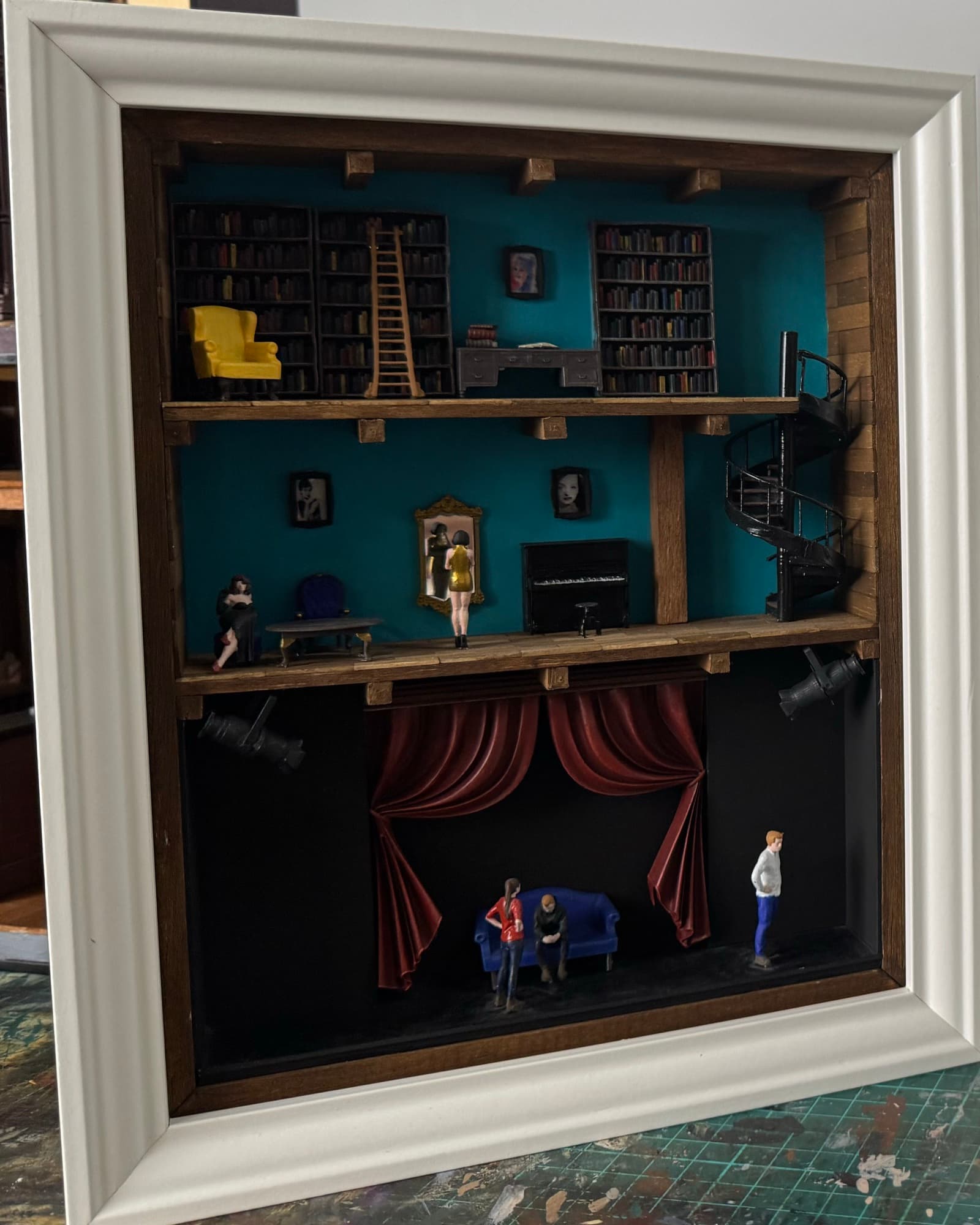 Framed miniature with a teal library, a spiral staircase and a theatre stage with red curtains