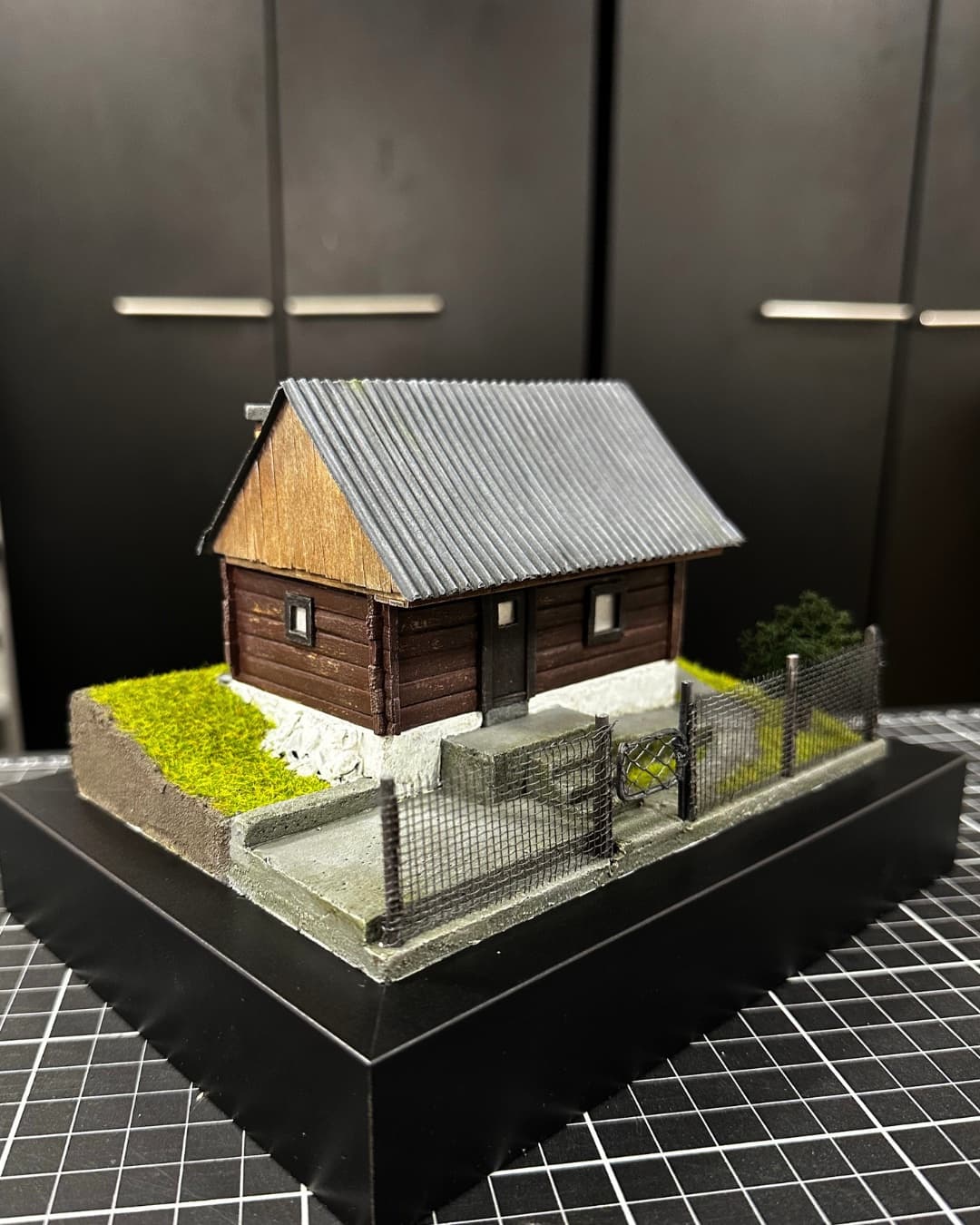 Diorama of a wooden log cabin with a metal roof and a fenced yard
