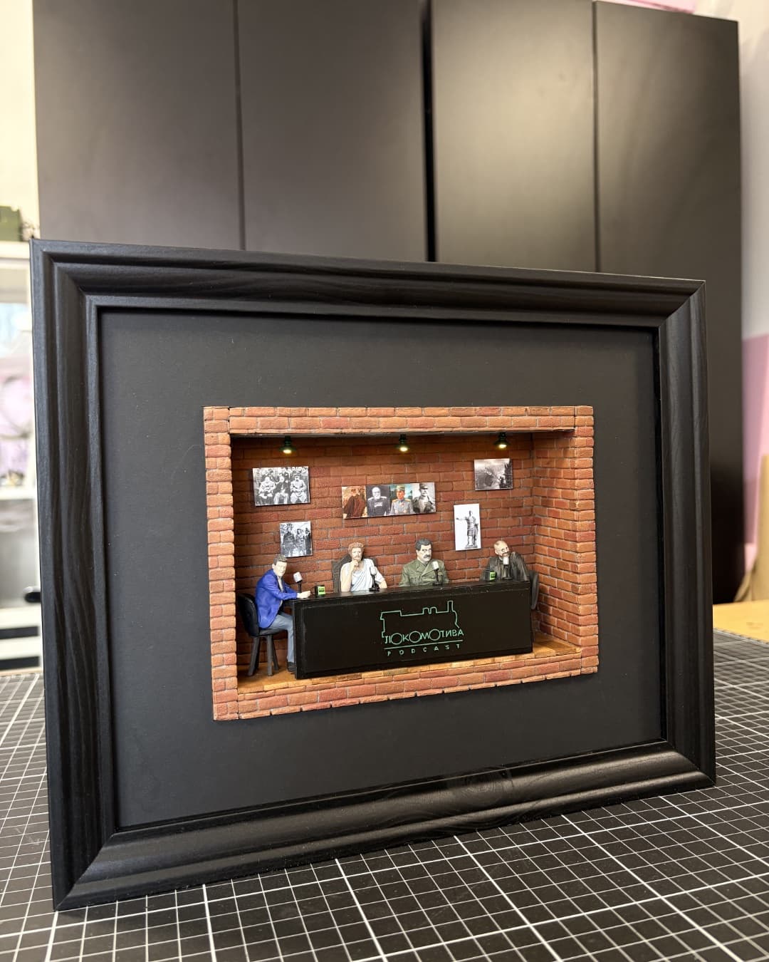 Framed miniature of a brick-walled podcast studio with four hosts at the desk